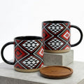 Two ceramic mugs with geometric patterns and a wooden coaster on a concrete surface.